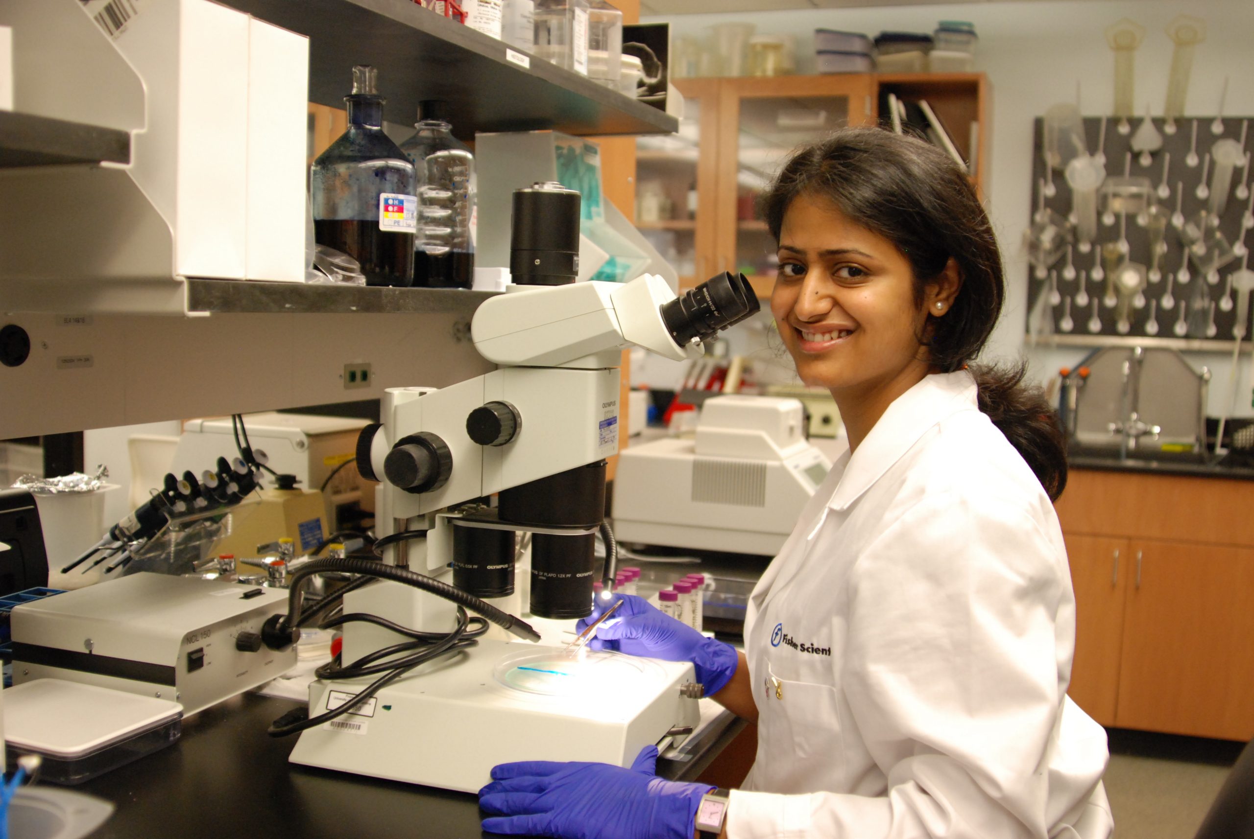 Woman in lab coat and gloves works at microscope in lab.